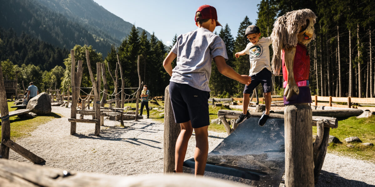 Zwei Kinder spielen an einem sonnigen Tag draußen auf einem Spielplatz mit Holzgeräten, umgeben von Bäumen und Bergen.