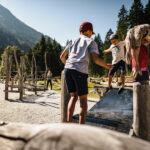 Zwei Kinder spielen an einem sonnigen Tag draußen auf einem Spielplatz mit Holzgeräten, umgeben von Bäumen und Bergen.