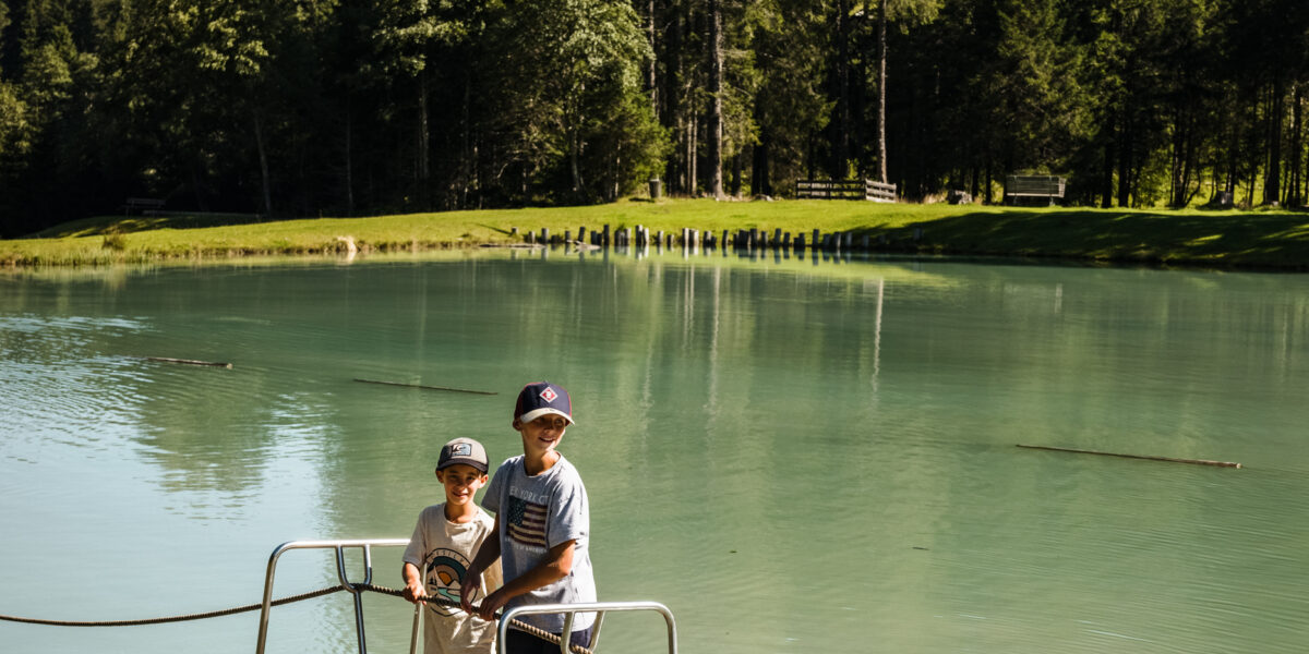Zwei Kinder stehen auf einem kleinen Holzfloß, das auf einem ruhigen See treibt, umgeben von hohen Bäumen und Bergen unter einem strahlend blauen Himmel.