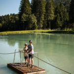 Zwei Kinder stehen auf einem kleinen Holzfloß, das auf einem ruhigen See treibt, umgeben von hohen Bäumen und Bergen unter einem strahlend blauen Himmel.