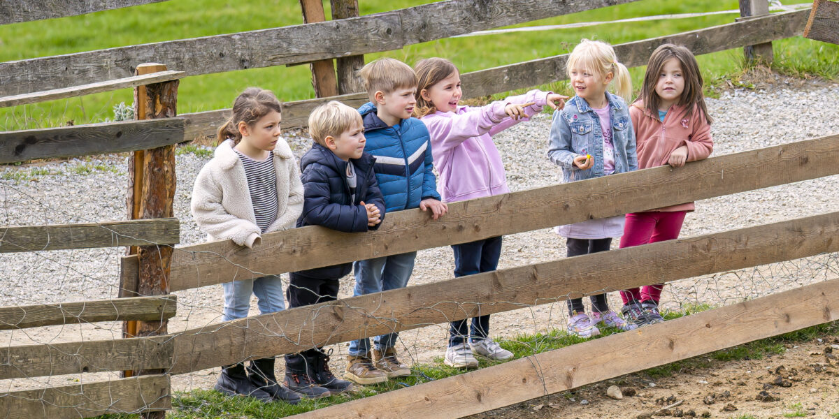 Sechs kleine Kinder stehen draußen an einem Holzzaun. Ein Kind zeigt nach vorne, während die anderen in dieselbe Richtung blicken. Im Hintergrund sind Gras und Kies zu sehen.