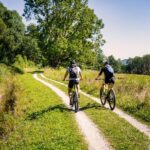 Zwei Personen fahren mit dem Fahrrad auf einem schmalen Feldweg durch eine grüne, bewaldete Landschaft unter einem klaren blauen Himmel.