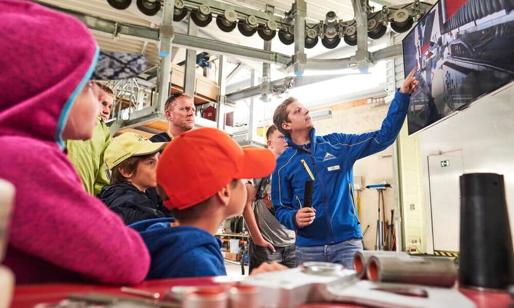 Eine Gruppe von Menschen, darunter Kinder und Erwachsene, beobachtet, wie ein Mann in einer blauen Jacke auf einen großen Bildschirm in einer Industriehalle zeigt.