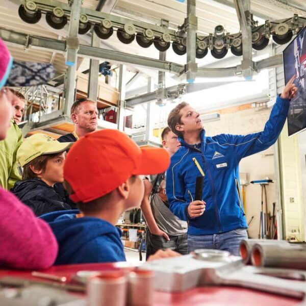 Eine Gruppe von Menschen, darunter Kinder und Erwachsene, beobachtet, wie ein Mann in einer blauen Jacke auf einen großen Bildschirm in einer Industriehalle zeigt.