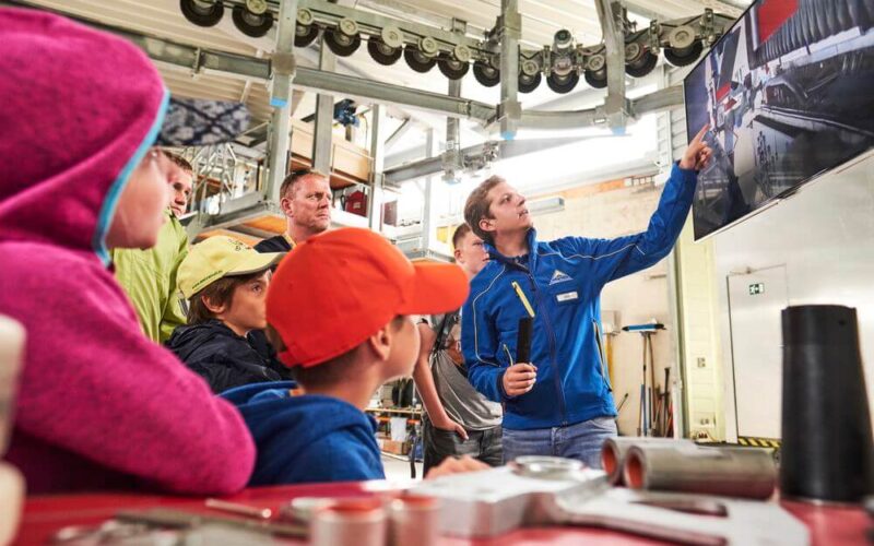 Eine Gruppe von Menschen, darunter Kinder und Erwachsene, beobachtet, wie ein Mann in einer blauen Jacke auf einen großen Bildschirm in einer Industriehalle zeigt.