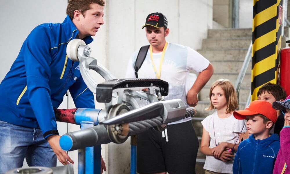 Ein Mann in einer blauen Jacke erklärt einer Gruppe von Kindern und einem Erwachsenen in einer Industrieumgebung ein mechanisches Gerät.