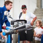 Ein Mann in einer blauen Jacke erklärt einer Gruppe von Kindern und einem Erwachsenen in einer Industrieumgebung ein mechanisches Gerät.