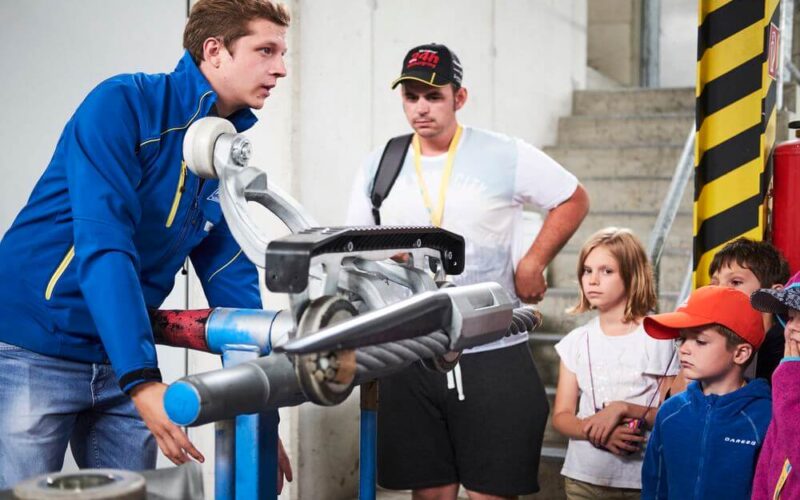 Ein Mann in einer blauen Jacke erklärt einer Gruppe von Kindern und einem Erwachsenen in einer Industrieumgebung ein mechanisches Gerät.