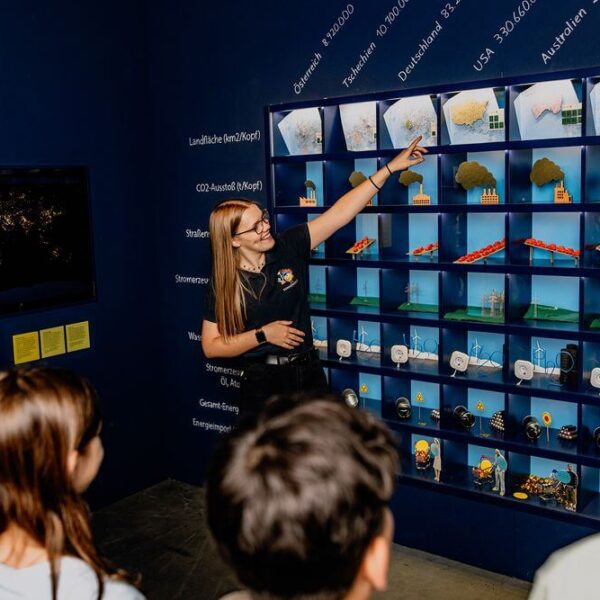 Eine Frau hält einen Vortrag vor einer Gruppe von Kindern und zeigt auf eine Tafel mit Ländernamen und verschiedenen Miniaturmodellen, die Umweltkonzepte darstellen.