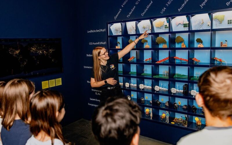 Eine Frau hält einen Vortrag vor einer Gruppe von Kindern und zeigt auf eine Tafel mit Ländernamen und verschiedenen Miniaturmodellen, die Umweltkonzepte darstellen.