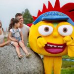 Ein Kind sitzt mit zwei Erwachsenen auf einem großen Felsen, während eine Person in einem leuchtend gelben Maskottchenkostüm mit der Aufschrift "Sonnenwelt Großschönau" neben ihnen im Freien posiert.