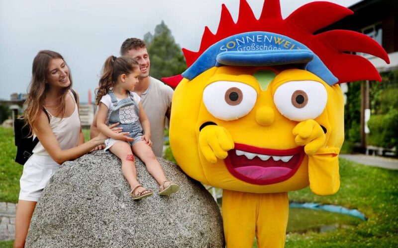 Ein Kind sitzt mit zwei Erwachsenen auf einem großen Felsen, während eine Person in einem leuchtend gelben Maskottchenkostüm mit der Aufschrift "Sonnenwelt Großschönau" neben ihnen im Freien posiert.