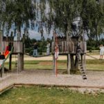 Die Kinder spielen auf einem hölzernen Spielplatz mit Rutschen, Leitern und einer Seilbrücke, umgeben von Bäumen und Wiesen.