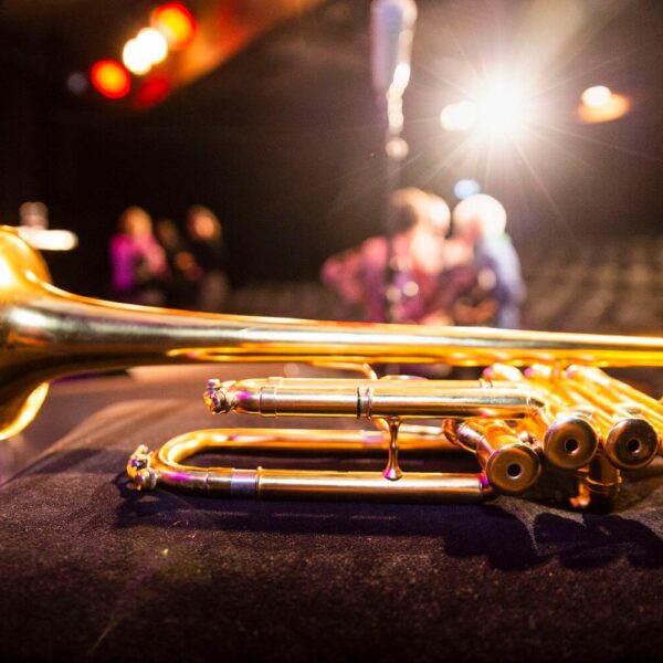 Nahaufnahme einer Messingtrompete, die auf einer dunklen Fläche ruht, mit Bühnenbeleuchtung und verschwommenen Menschen im Hintergrund.