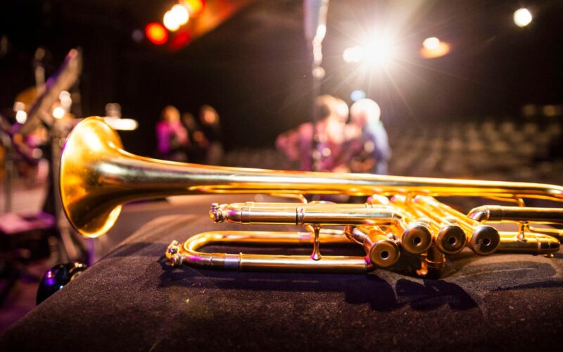 Nahaufnahme einer Messingtrompete, die auf einer dunklen Fläche ruht, mit Bühnenbeleuchtung und verschwommenen Menschen im Hintergrund.