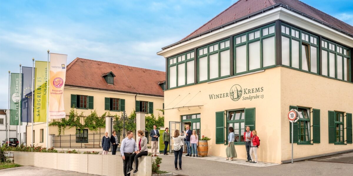 Menschen stehen und gehen vor dem Gebäude Winzer Krems Sandgrube 13, das eine beige Fassade mit grüner Umrandung hat und in dessen Nähe unter einem teilweise bewölkten Himmel mehrere Fahnen wehen.
