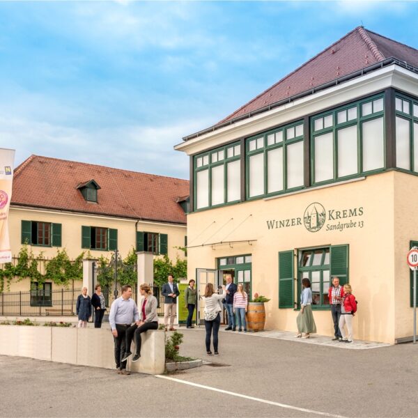 Menschen stehen und gehen vor dem Gebäude Winzer Krems Sandgrube 13, das eine beige Fassade mit grüner Umrandung hat und in dessen Nähe unter einem teilweise bewölkten Himmel mehrere Fahnen wehen.