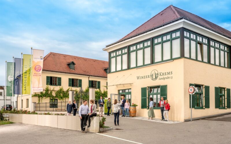 Menschen stehen und gehen vor dem Gebäude Winzer Krems Sandgrube 13, das eine beige Fassade mit grüner Umrandung hat und in dessen Nähe unter einem teilweise bewölkten Himmel mehrere Fahnen wehen.