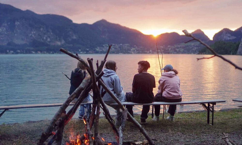 Vier Personen sitzen bei Sonnenuntergang auf einer Bank an einem See, mit Bergen im Hintergrund und einem brennenden Lagerfeuer im Vordergrund.
