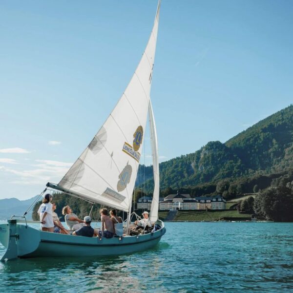 Eine Gruppe von Menschen fährt mit einem Segelboot auf einem ruhigen See mit bewaldeten Hügeln und Gebäuden im Hintergrund unter einem klaren Himmel.