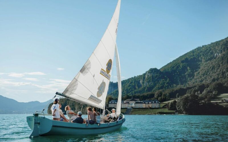 Eine Gruppe von Menschen fährt mit einem Segelboot auf einem ruhigen See mit bewaldeten Hügeln und Gebäuden im Hintergrund unter einem klaren Himmel.