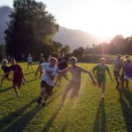 Eine Gruppe von Kindern und Jugendlichen rennt und spielt auf einer Wiese in hellem Sonnenlicht mit Bäumen und Bergen im Hintergrund.