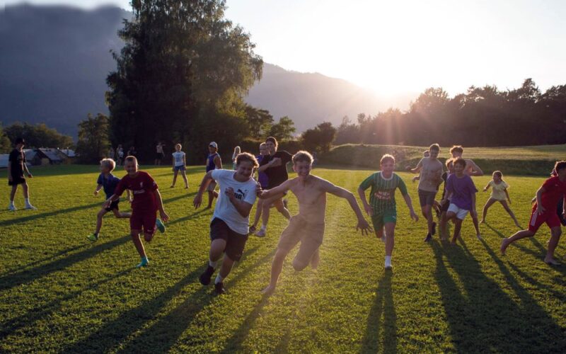 Eine Gruppe von Kindern und Jugendlichen rennt und spielt auf einer Wiese in hellem Sonnenlicht mit Bäumen und Bergen im Hintergrund.