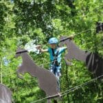 Ein Kind in Sicherheitsausrüstung balanciert auf einem Hochseilgarten und läuft über pferdeförmige Metallplattformen, die zwischen Bäumen in einem Waldgebiet aufgehängt sind.