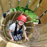 Ein Mädchen, das einen Helm und einen Klettergurt trägt, krabbelt durch einen Holztunnel in einem Outdoor-Abenteuerpark.