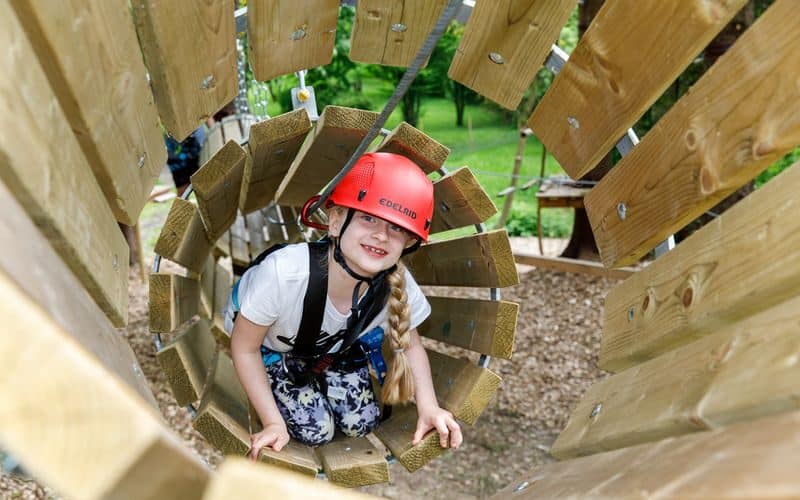 Ein Mädchen, das einen Helm und einen Klettergurt trägt, krabbelt durch einen Holztunnel in einem Outdoor-Abenteuerpark.