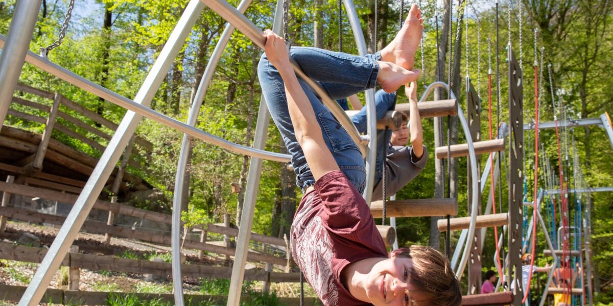 Ein Kind hängt kopfüber an den Beinen von einem Spielplatzgerät und lächelt, während im Hintergrund Bäume und andere Spielplatzstrukturen zu sehen sind.