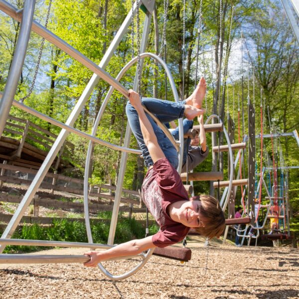 Ein Kind hängt kopfüber an den Beinen von einem Spielplatzgerät und lächelt, während im Hintergrund Bäume und andere Spielplatzstrukturen zu sehen sind.