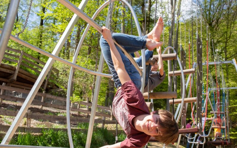 Ein Kind hängt kopfüber an den Beinen von einem Spielplatzgerät und lächelt, während im Hintergrund Bäume und andere Spielplatzstrukturen zu sehen sind.
