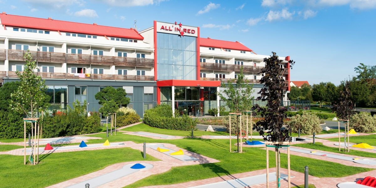 Ein modernes Hotel mit rotem Dach und der Aufschrift "All in Red" steht hinter einem begrünten Minigolfplatz unter einem teilweise bewölkten Himmel.