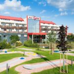 Ein modernes Hotel mit rotem Dach und der Aufschrift "All in Red" steht hinter einem begrünten Minigolfplatz unter einem teilweise bewölkten Himmel.