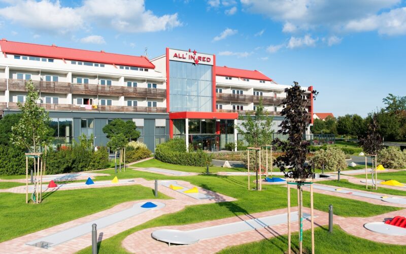 Ein modernes Hotel mit rotem Dach und der Aufschrift "All in Red" steht hinter einem begrünten Minigolfplatz unter einem teilweise bewölkten Himmel.