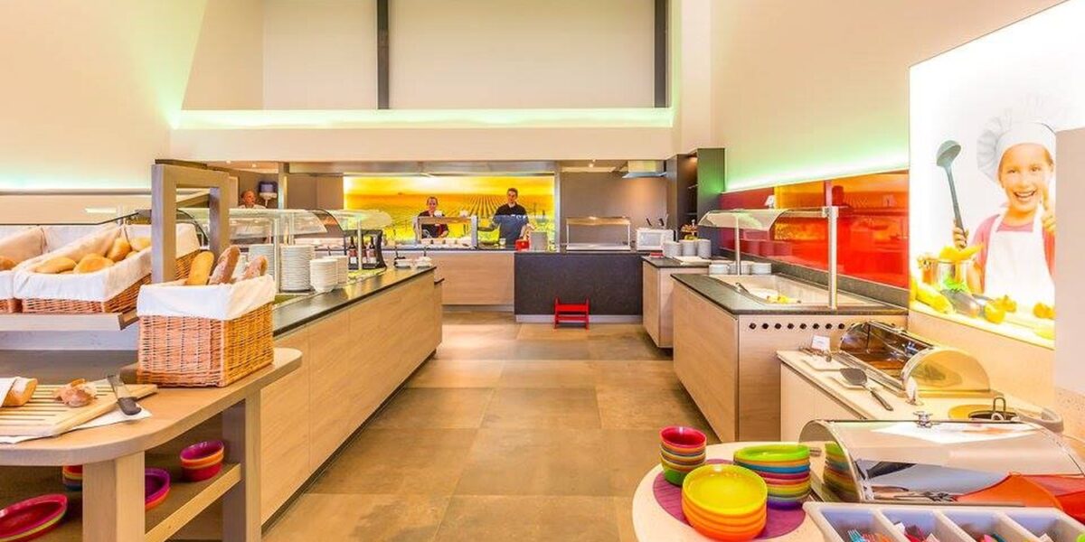 Eine hell erleuchtete Selbstbedienungs-Cafeteria mit einer Vielzahl von Essensstationen, bunten Schüsseln, Brotkörben und zwei Mitarbeitern hinter der Theke.