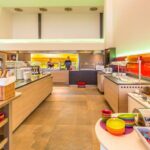 Eine hell erleuchtete Selbstbedienungs-Cafeteria mit einer Vielzahl von Essensstationen, bunten Schüsseln, Brotkörben und zwei Mitarbeitern hinter der Theke.