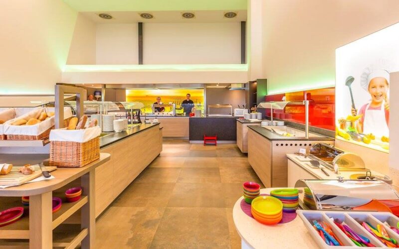 Eine hell erleuchtete Selbstbedienungs-Cafeteria mit einer Vielzahl von Essensstationen, bunten Schüsseln, Brotkörben und zwei Mitarbeitern hinter der Theke.