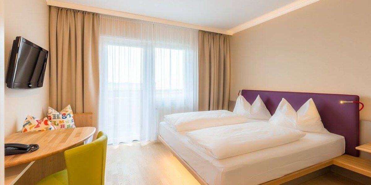 Ein modernes Hotelzimmer mit einem Doppelbett, einem kleinen Schreibtisch und einem grünen Stuhl, einem an der Wand montierten Fernseher, einem großen Fenster mit Vorhängen und hellem Holzfußboden.