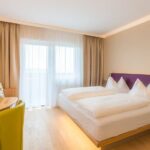 Ein modernes Hotelzimmer mit einem Doppelbett, einem kleinen Schreibtisch und einem grünen Stuhl, einem an der Wand montierten Fernseher, einem großen Fenster mit Vorhängen und hellem Holzfußboden.