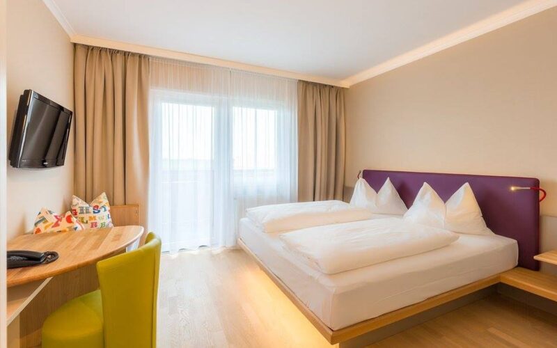 Ein modernes Hotelzimmer mit einem Doppelbett, einem kleinen Schreibtisch und einem grünen Stuhl, einem an der Wand montierten Fernseher, einem großen Fenster mit Vorhängen und hellem Holzfußboden.