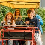 Vier Personen sitzen zusammen auf einem Vierrad mit zwei Lenkrädern, lächeln in die Kamera und sind von einer grünen Landschaft umgeben.