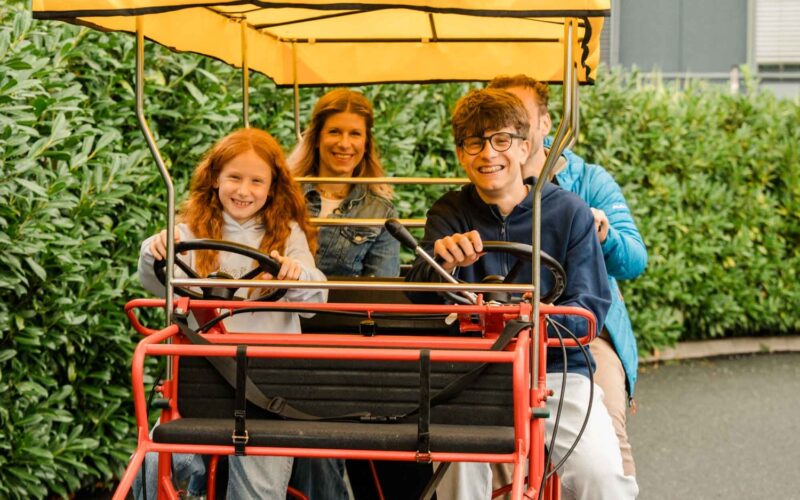 Vier Personen sitzen zusammen auf einem Vierrad mit zwei Lenkrädern, lächeln in die Kamera und sind von einer grünen Landschaft umgeben.