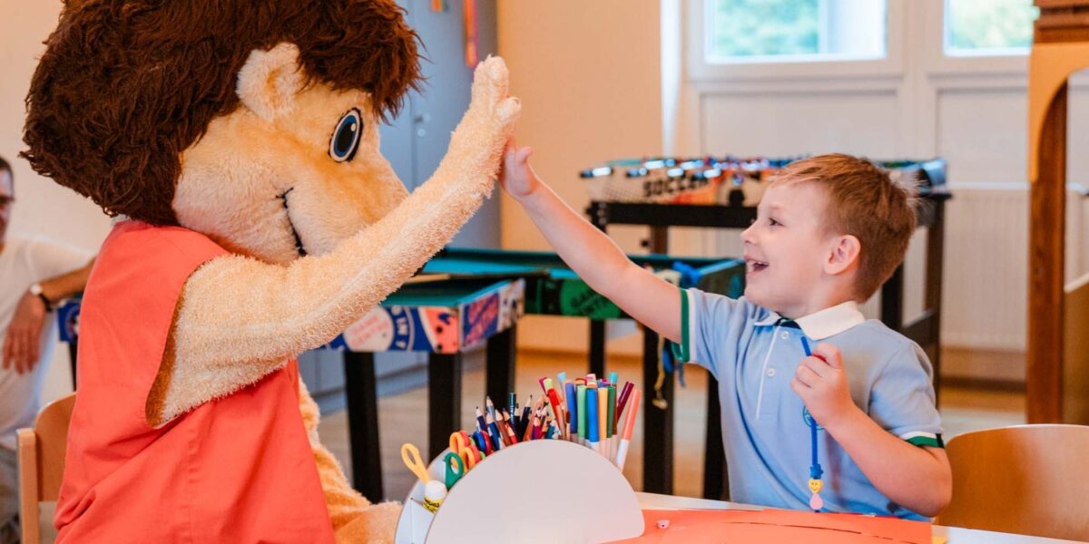 Ein Kind, das an einem Basteltisch sitzt, gibt einer Person in einem großen Tierkostüm in einem hell erleuchteten Klassenzimmer ein High-Five.