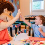 Ein Kind, das an einem Basteltisch sitzt, gibt einer Person in einem großen Tierkostüm in einem hell erleuchteten Klassenzimmer ein High-Five.