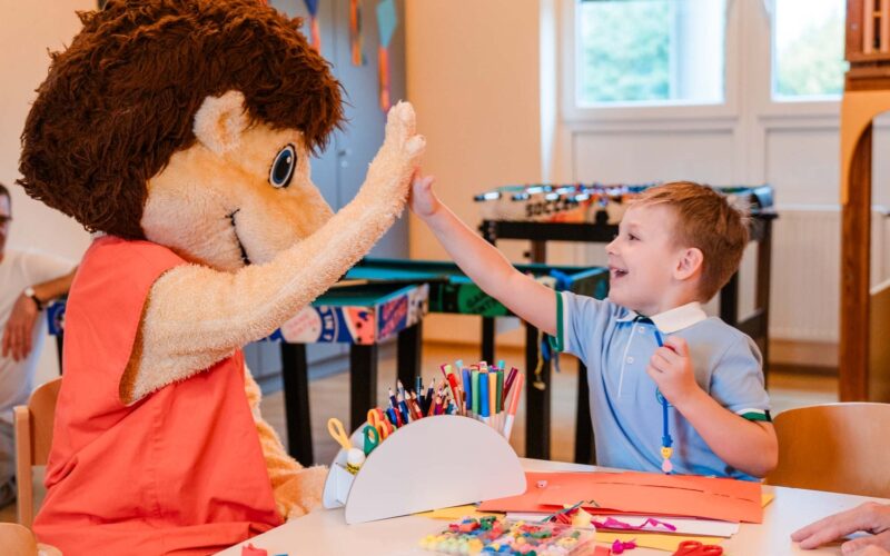 Ein Kind, das an einem Basteltisch sitzt, gibt einer Person in einem großen Tierkostüm in einem hell erleuchteten Klassenzimmer ein High-Five.