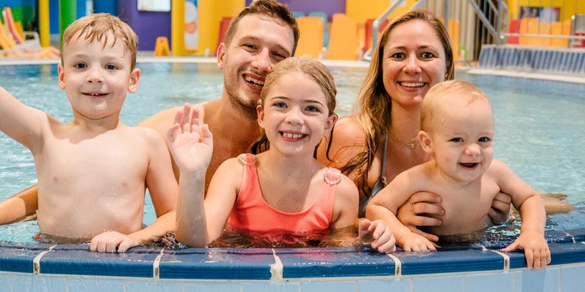 Eine lächelnde Familie mit zwei Erwachsenen und drei Kindern posiert gemeinsam im flachen Ende eines Hallenbades.