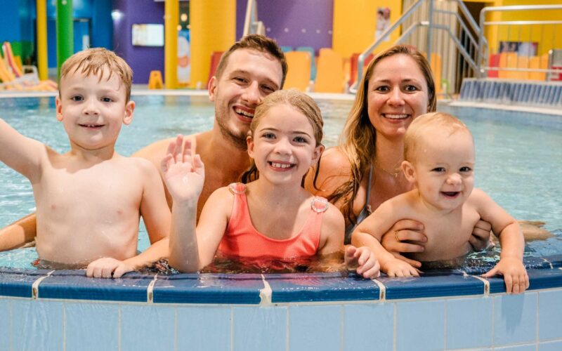 Eine lächelnde Familie mit zwei Erwachsenen und drei Kindern posiert gemeinsam im flachen Ende eines Hallenbades.