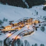 Luftaufnahme eines schneebedeckten Bergdorfs in der Abenddämmerung mit warm beleuchteten Gebäuden, umgeben von Bäumen und Hügeln.